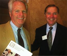 Two Men in Suits, Smiling and Standing Together, One of Them Holding a Branded Paper at an Event