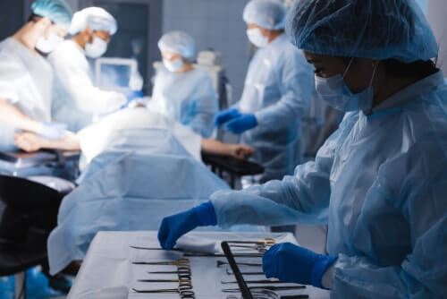Surgeon Arranging Tools With Operating Table With Patient Laying in it Surrounded by Surgeons in the Background