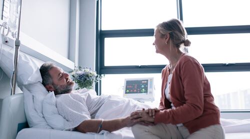 Man Laying in Hospital Bed Holding Woman's Hand As She Sits on the Side of the Bed